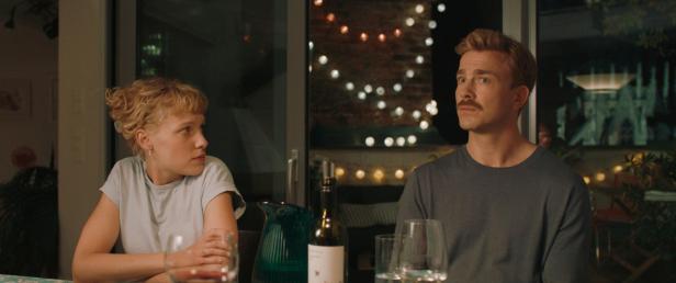 Ein Paar sitzt an einem Tisch mit Wein und Gläsern, im Hintergrund Lichterketten.