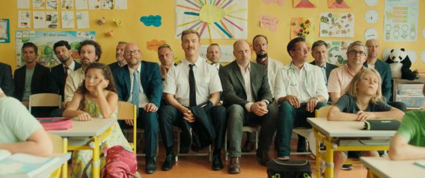 Eine Gruppe von Erwachsenen sitzt in einem Klassenzimmer, einige Kinder sitzen an Schreibtischen.