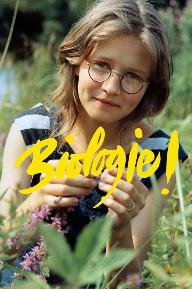 Eine junge Frau mit Brille betrachtet eine kleine Blume in einer Wiese.