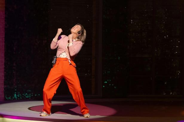 Renée Zellweger in orangefarbener Hose und rosa Strickjacke auf einer Bühne.