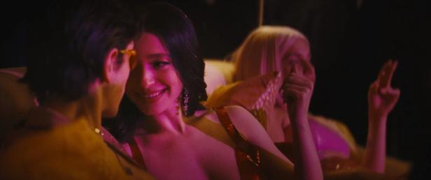 Eine Frau mit Ohrringen lächelt einen Mann an, während eine andere Frau im Hintergrund ein Victory-Zeichen macht.