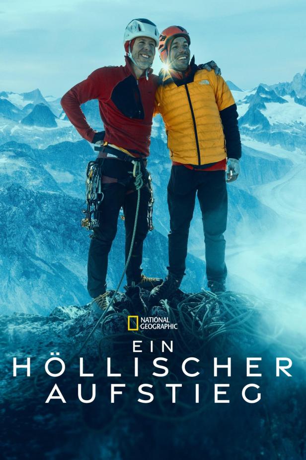 Zwei Bergsteiger stehen auf einem Gipfel mit schneebedeckten Bergen im Hintergrund.