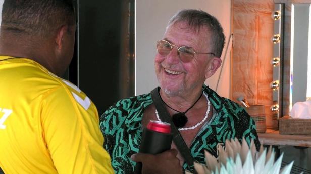 Ein lächelnder älterer Mann mit Brille und offenem Hemd hält ein Getränk in der Hand und unterhält sich mit jemandem.