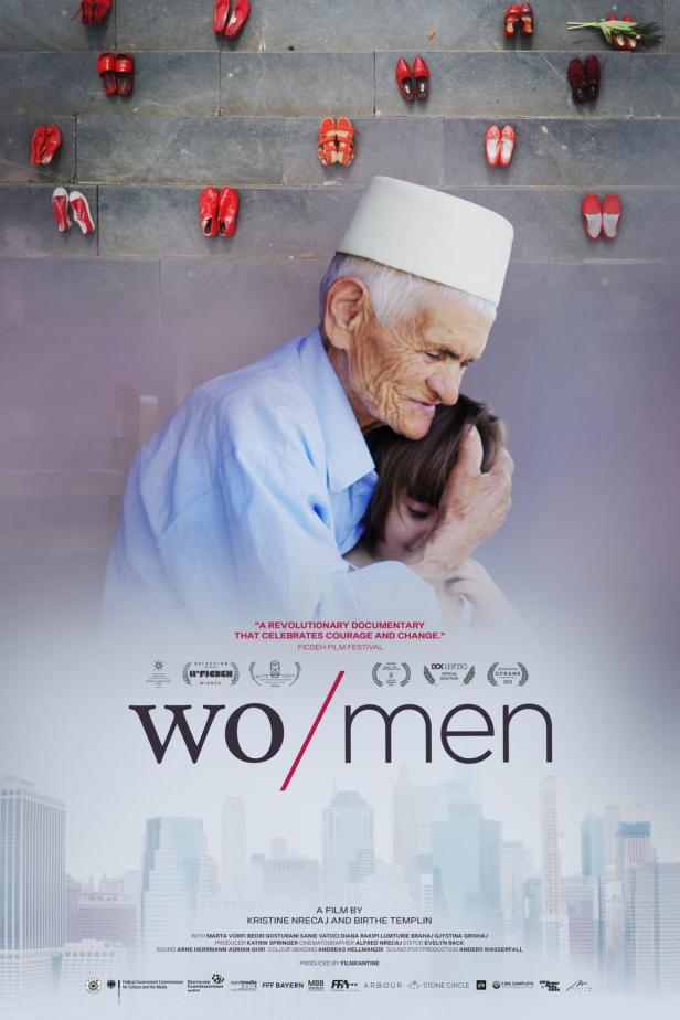 Ein älterer Mann mit traditioneller Kopfbedeckung umarmt ein junges Mädchen vor dem Hintergrund des Filmplakats „Wo/men“.