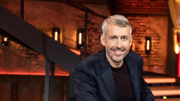 Ein Mann mit grauem Haar und Bart lächelt in die Kamera, im Hintergrund eine Treppe.