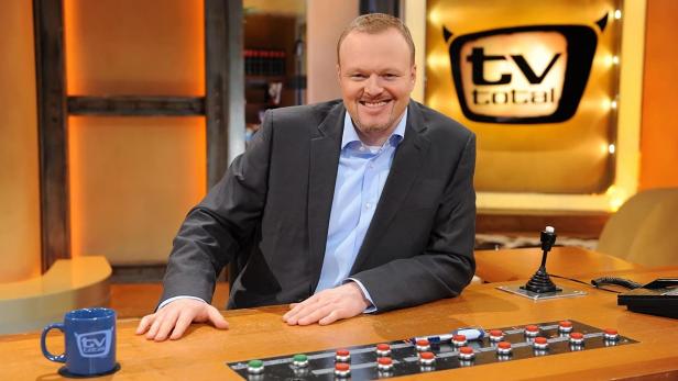 Stefan Raab sitzt lächelnd am Schreibtisch von „TV total“.
