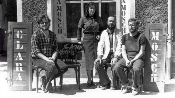 Eine Gruppe von vier Personen sitzt vor einem Gebäude mit der Aufschrift „Amosch“ und „Clara“.