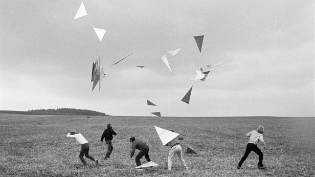 Fünf Personen rennen auf einem Feld unter einem Schwarm dreieckiger Drachen umher.