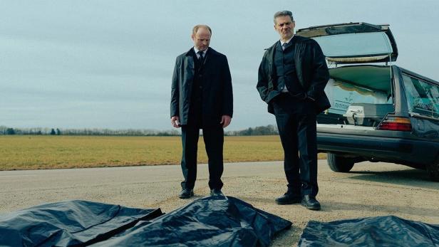 Zwei Männer in Anzügen stehen vor Leichenwagen und abgedeckten Leichen.