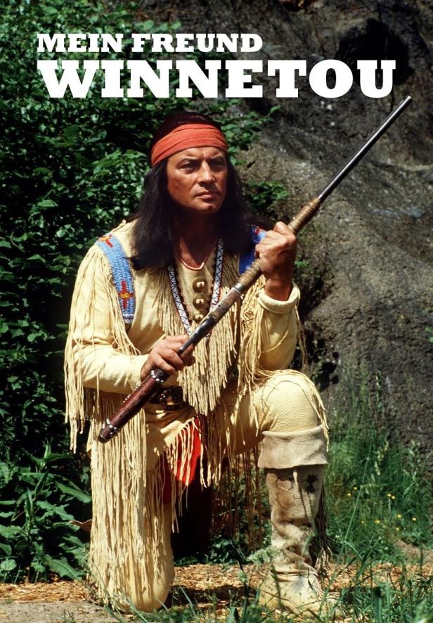 Ein Mann in traditioneller Winnetou-Kleidung kniet mit einem Gewehr in der Hand.
