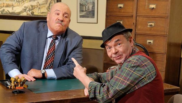 Zwei Männer sitzen an einem Schreibtisch, einer zeigt mit dem Finger auf etwas.