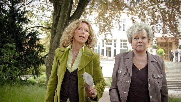 Zwei Frauen gehen in einem Park an einem Gebäude vorbei, eine hält einen Schuh in der Hand.