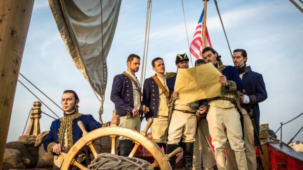 Eine Gruppe von Männern in historischen Marineuniformen studiert eine Karte auf einem Segelschiff.