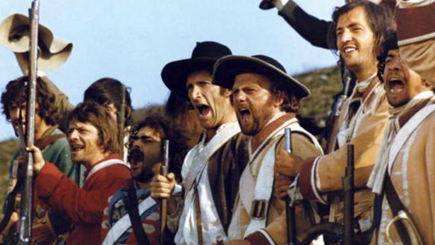 Eine Gruppe von Männern in historischen Kostümen schreit und hält Gewehre.