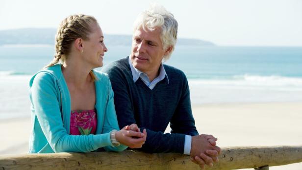 Eine junge Frau und ein älterer Mann lehnen an einer hölzernen Barriere am Strand.