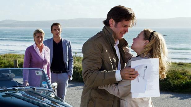 Ein Paar umarmt sich innig vor einer Küstenlandschaft, während zwei weitere Personen im Hintergrund stehen.