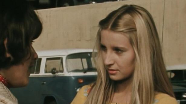 Eine junge Frau mit blonden Haaren steht vor einem blauen VW-Bus.