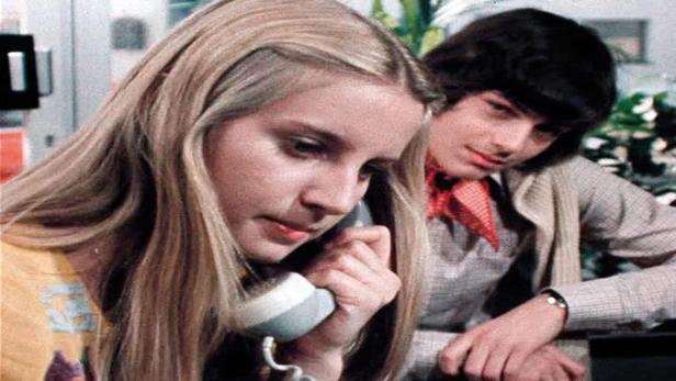 Eine junge Frau telefoniert, während ein junger Mann ihr über die Schulter schaut.