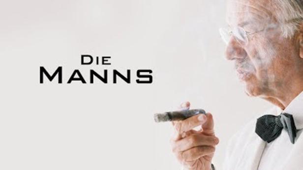 Ein älterer Mann mit Fliege raucht eine Zigarre; im Hintergrund der Schriftzug „Die Manns“.