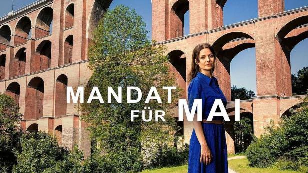 Eine Frau steht vor einer alten Ziegelsteinbrücke; Wahlwerbung „Mandat für Mai“.