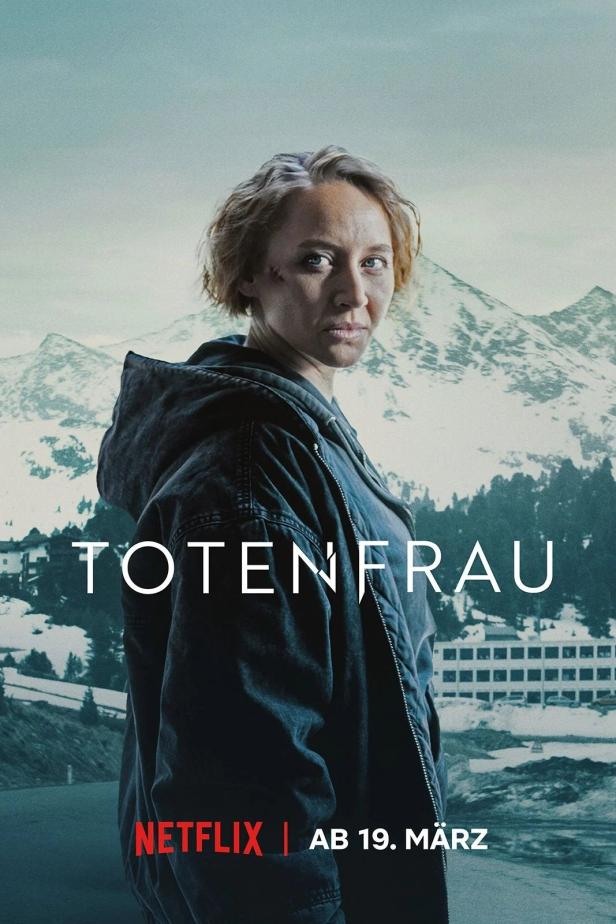 Eine Frau mit einer Narbe im Gesicht steht vor einer verschneiten Berglandschaft.