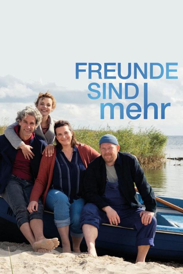 Vier Freunde sitzen barfuß am Strand vor einem See, im Hintergrund der Titel „Freunde sind mehr“.