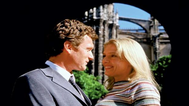 Ein Mann im Anzug und eine blonde Frau schauen sich vor einer Kathedrale anlächelnd an.