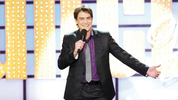 Ein Mann im Anzug steht auf einer Bühne und spricht in ein Mikrofon.