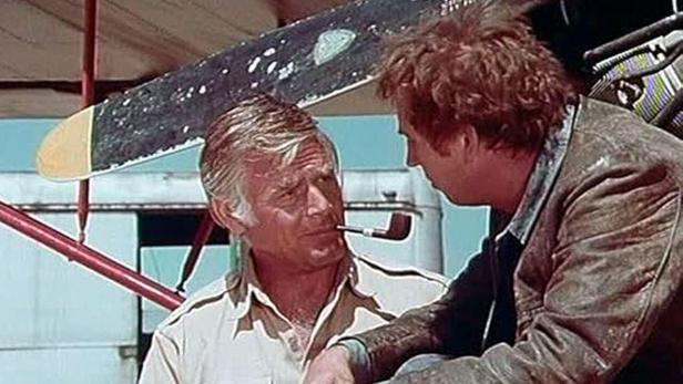 Zwei Männer unterhalten sich vor einem Flugzeug, einer raucht Pfeife.