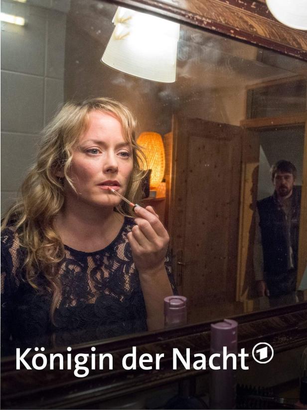 Eine Frau trägt Lippenstift vor einem Spiegel auf, im Hintergrund ein Mann.