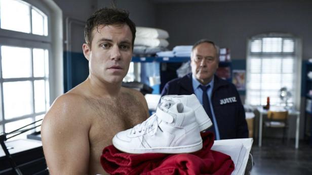 Ein verschwitzter, barbusiger Mann hält ein Paar weiße Turnschuhe und ein rotes T-Shirt.