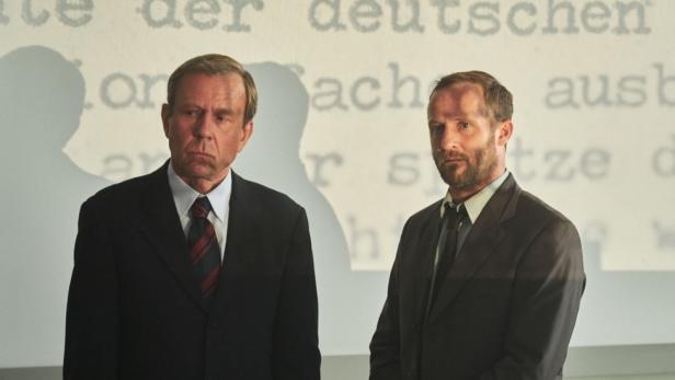 Zwei Männer in Anzügen stehen vor einer Wand mit Textfragmenten.