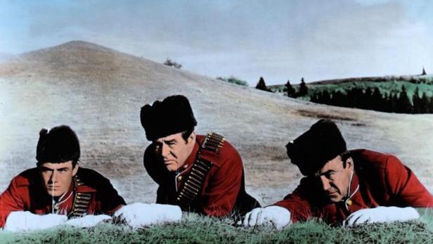 Drei Männer in roten Uniformen und Pelzmützen liegen im Gras vor einer Hügellandschaft.