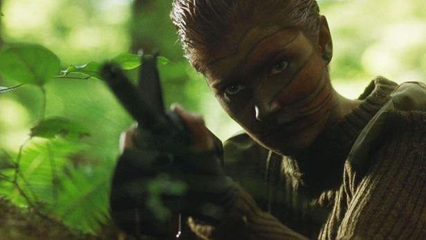 Eine Soldatin mit Gesichtsbemalung zielt mit einer Waffe im Grünen.