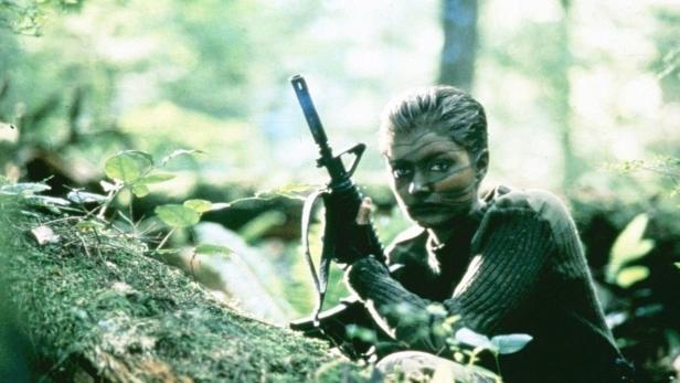 Eine Soldatin mit Gesichtsbemalung hält ein Gewehr im Wald.