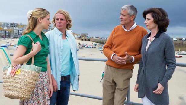 Zwei Paare unterhalten sich an einem Strand mit einem kleinen Fischerdorf im Hintergrund.