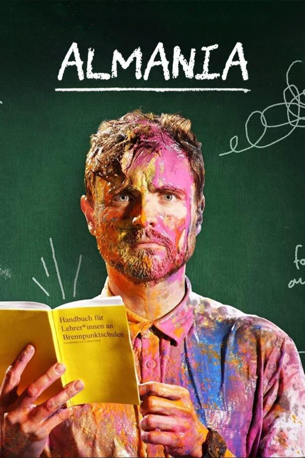 Ein Mann, mit Farbe beschmiert, hält ein Handbuch für Lehrer an Brennpunktschulen vor einer Tafel mit dem Wort „Almania“.