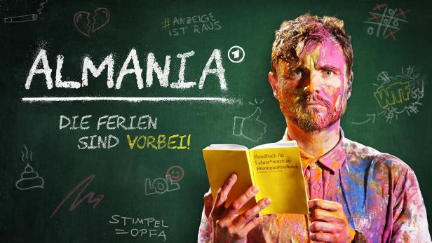 Ein mit Farbe beschmierter Mann hält ein Buch mit dem Titel „Handbuch für Lehrer*innen an Brennpunktschulen“ vor einer Tafel mit der Aufschrift „Almania“.