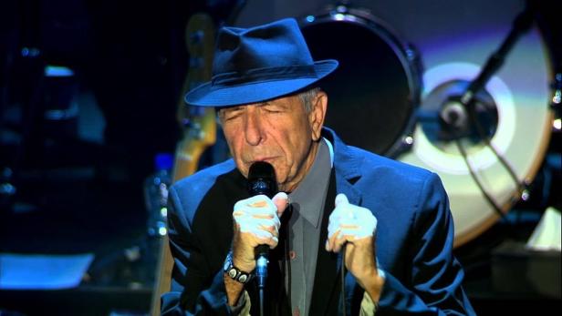 Leonard Cohen singt mit Hut und Anzug auf einer Bühne.