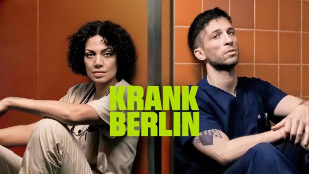 Zwei junge Pflegekräfte sitzen vor orangefarbenen Kacheln, im Vordergrund der Schriftzug „Krank Berlin“.