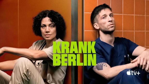 Zwei medizinische Fachkräfte, vermutlich aus der Serie „Krank Berlin“, sitzen vor orangefarbenen Wänden.