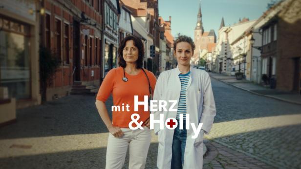 Zwei Ärztinnen stehen vor einer Altstadtkulisse, im Vordergrund der Schriftzug „mit Herz & Holly“.