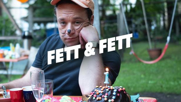 Ein Mann mit Katzengesicht sitzt traurig an einem Geburtstagstisch mit der Aufschrift „Fett & Fett“.