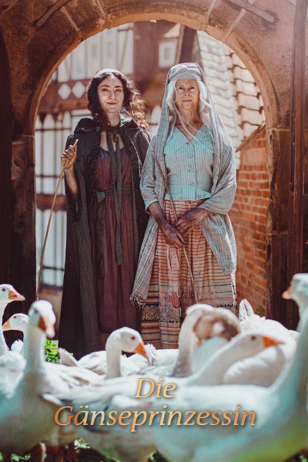Zwei Frauen stehen vor einem Torbogen, umgeben von Gänsen, im Film „Die Gänseprinzessin“.