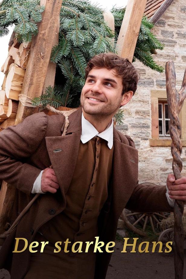 Ein lächelnder Mann in historischer Kleidung trägt Holz und Tannengrün vor einem Haus.