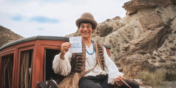 Ein Mann im Kostüm sitzt auf einer Kutsche und hält eine Ankündigung für eine Neueröffnung in der Hand.