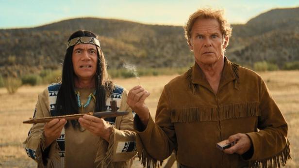 Zwei Männer in Kostümen stehen in einer Prärielandschaft, einer zündet ein Streichholz an.