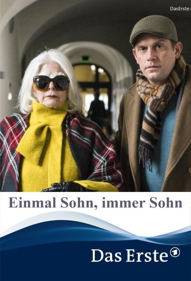 Eine ältere Frau mit Sonnenbrille und ein Mann mit Schal stehen nebeneinander.