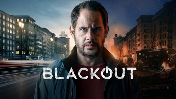 Ein Mann vor dem Hintergrund des Posters für die Fernsehserie „Blackout“.