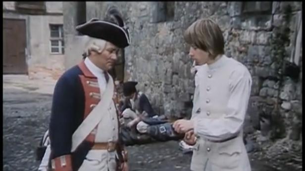 Zwei Männer in historischen Kostümen unterhalten sich vor einer Steinmauer.